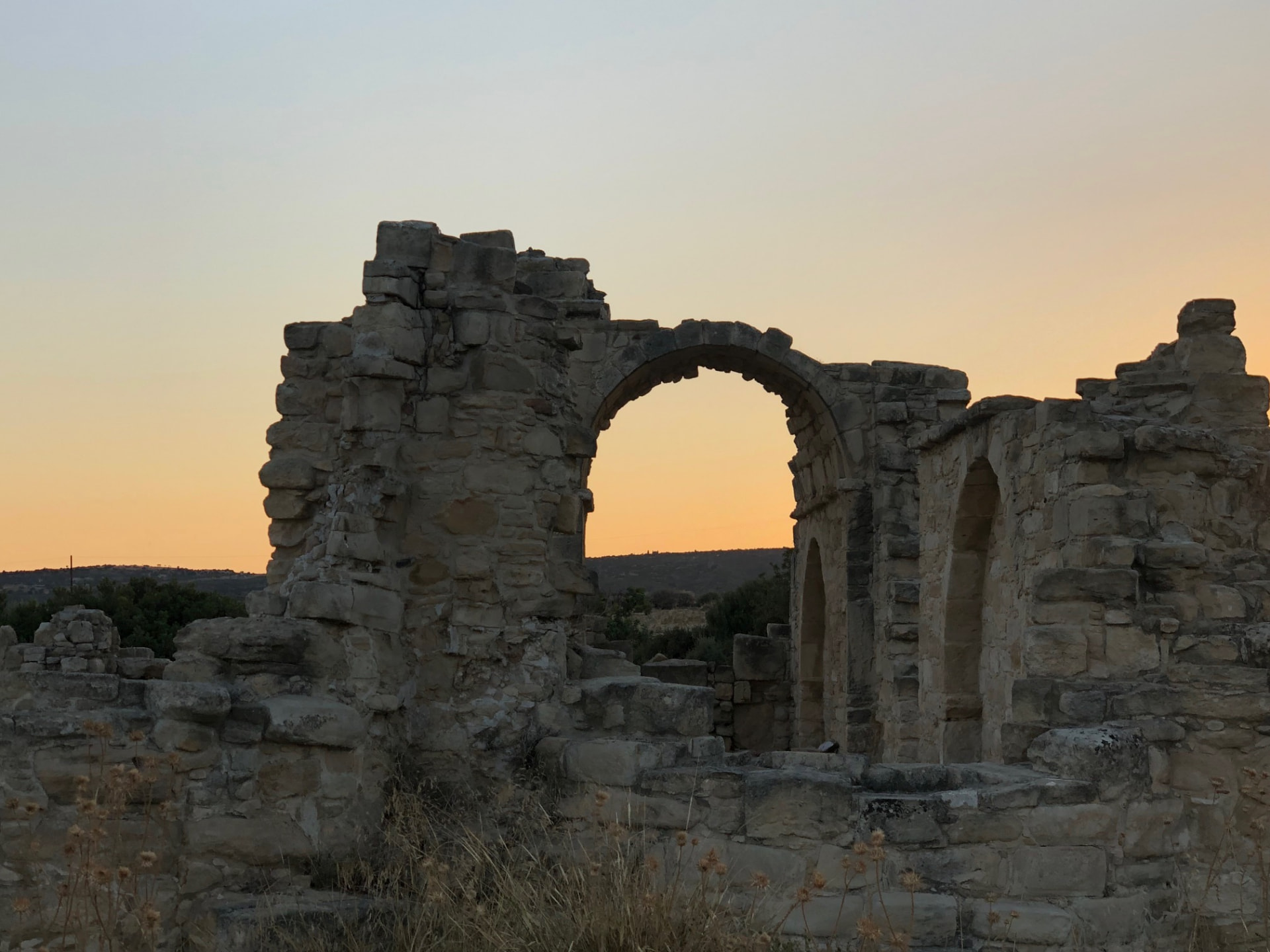 Ruins of Agios Tychikos at sunset, with a stone arch framing the golden sky over Palodeia village