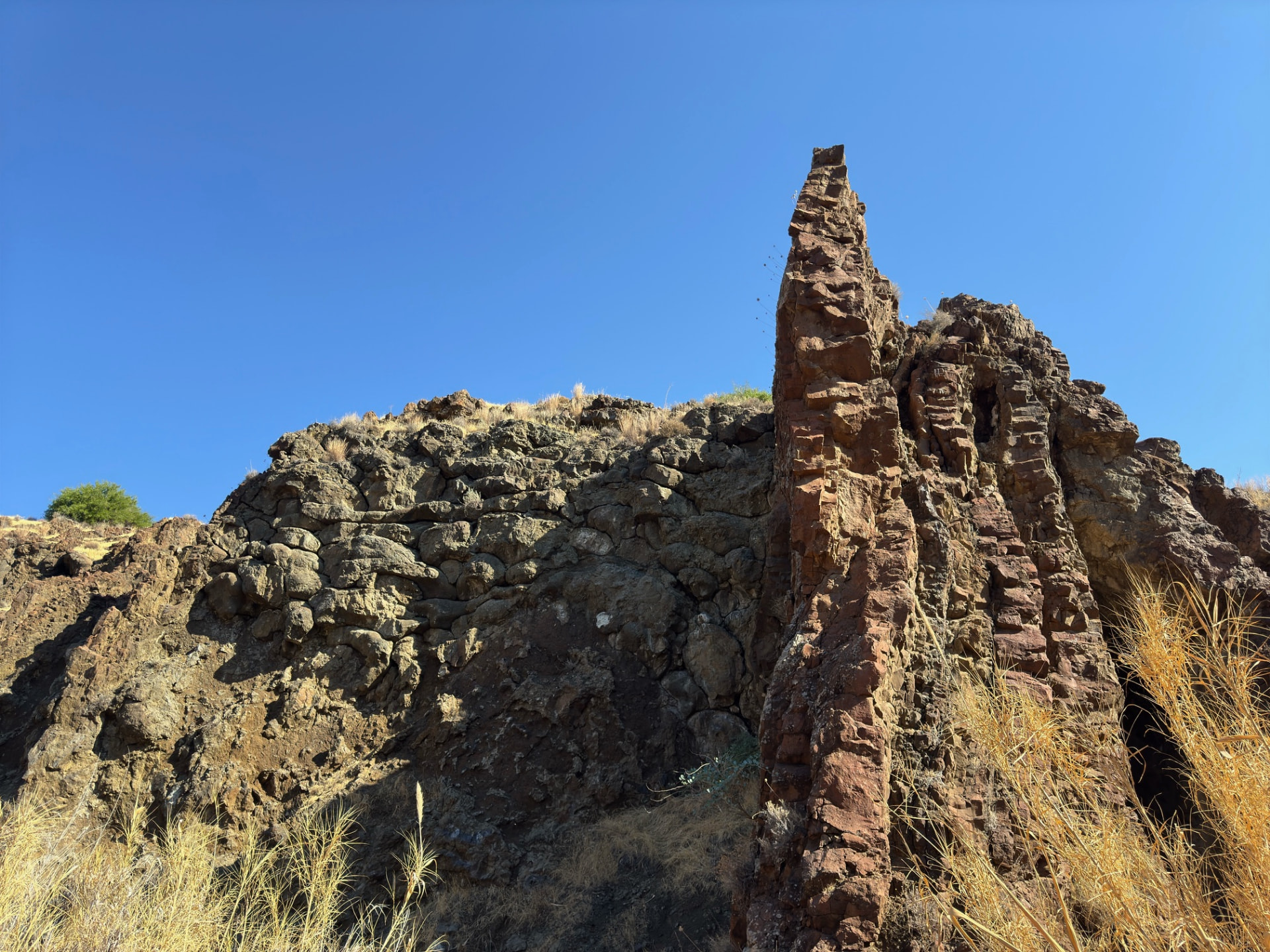 Jagged volcanic rock formation rising from dry grass under a clear blue sky at Pikrovrisi tis Merikas